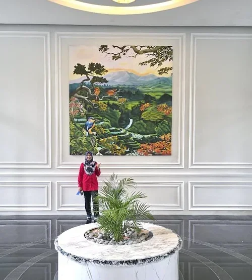 A woman in a pink jacket and scarf standing in front of a large landscape painting inside a building.