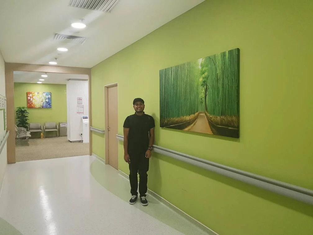 A young man standing in a hospital corridor with light green walls, a large bamboo forest painting, and a woman sitting in the background.