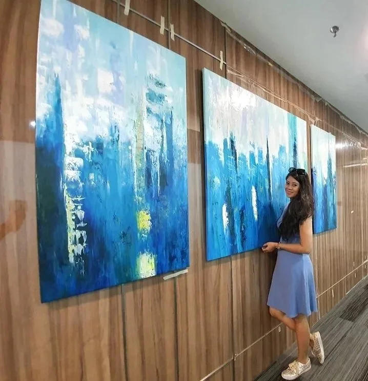 A young woman in a blue dress and white sneakers standing and looking at three large blue abstract paintings hung on a wood-paneled wall.