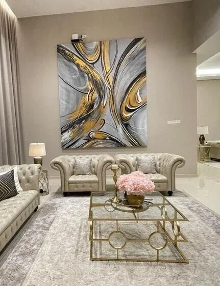 Modern living room with beige tufted sofas, a glass and gold coffee table with pink flowers, abstract wall art in gray, white, and gold, soft lighting, and neutral decor.