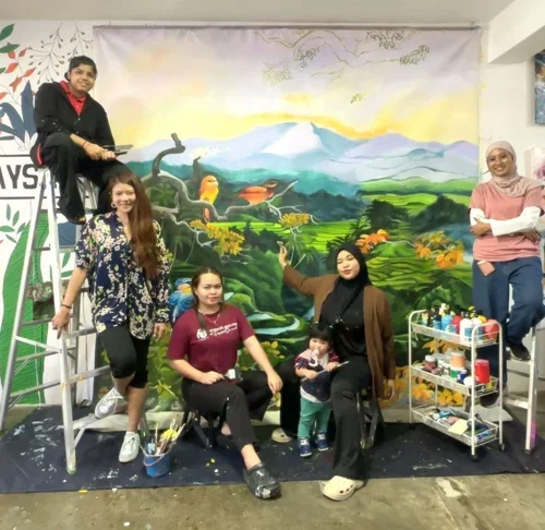 Group of five women and a young girl in front of a large painted mural with a landscape scene. Two women stand on ladders, one on the ground, and one woman is sitting with a child. Art supplies are on a cart nearby.