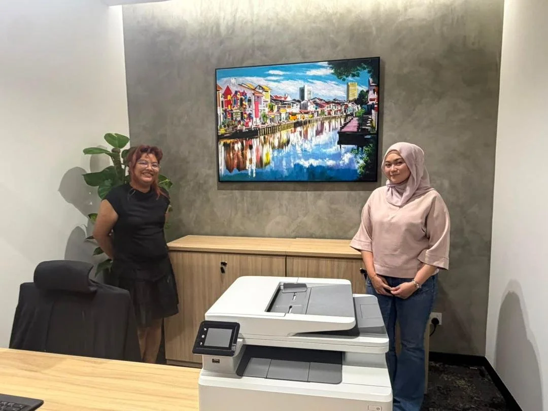 Two women stand in an office with a large colorful cityscape painting on the wall behind them and a machine on the table in front of them.
