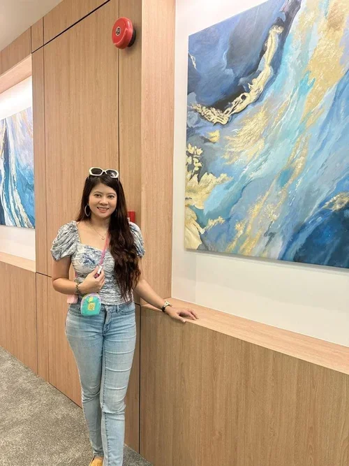A young woman with long dark hair in a floral top and jeans stands in an art gallery, smiling at the camera. She has sunglasses on her head, a small crossbody bag, and is standing next to a large abstract painting with blue, yellow, and white colors.