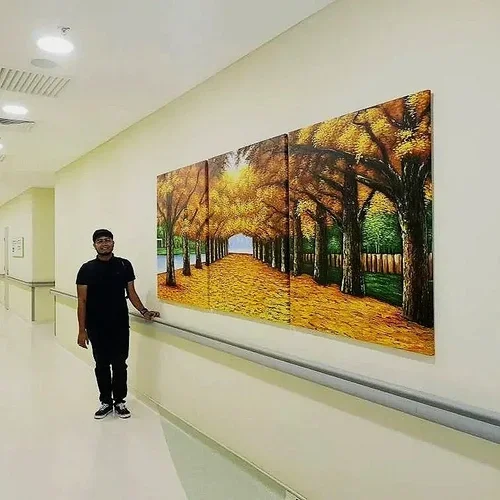 A man standing in a hallway next to a large, four-panel landscape painting of an autumn scene with yellow and orange trees and a pathway, on a white wall.