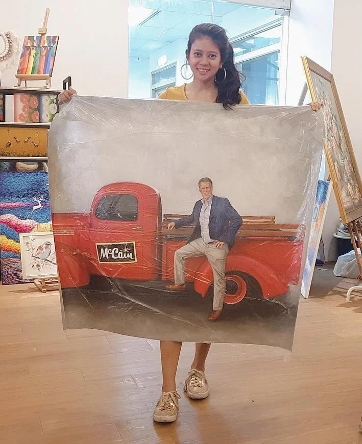 Young girl holding a large printed photo of a man in a suit standing next to a vintage red truck with the name 'McCain' on the door, inside an art gallery.