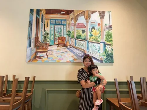 A woman holding a young child in a dining area with wooden chairs, standing in front of a large colorful painting of a veranda with plants and trees.