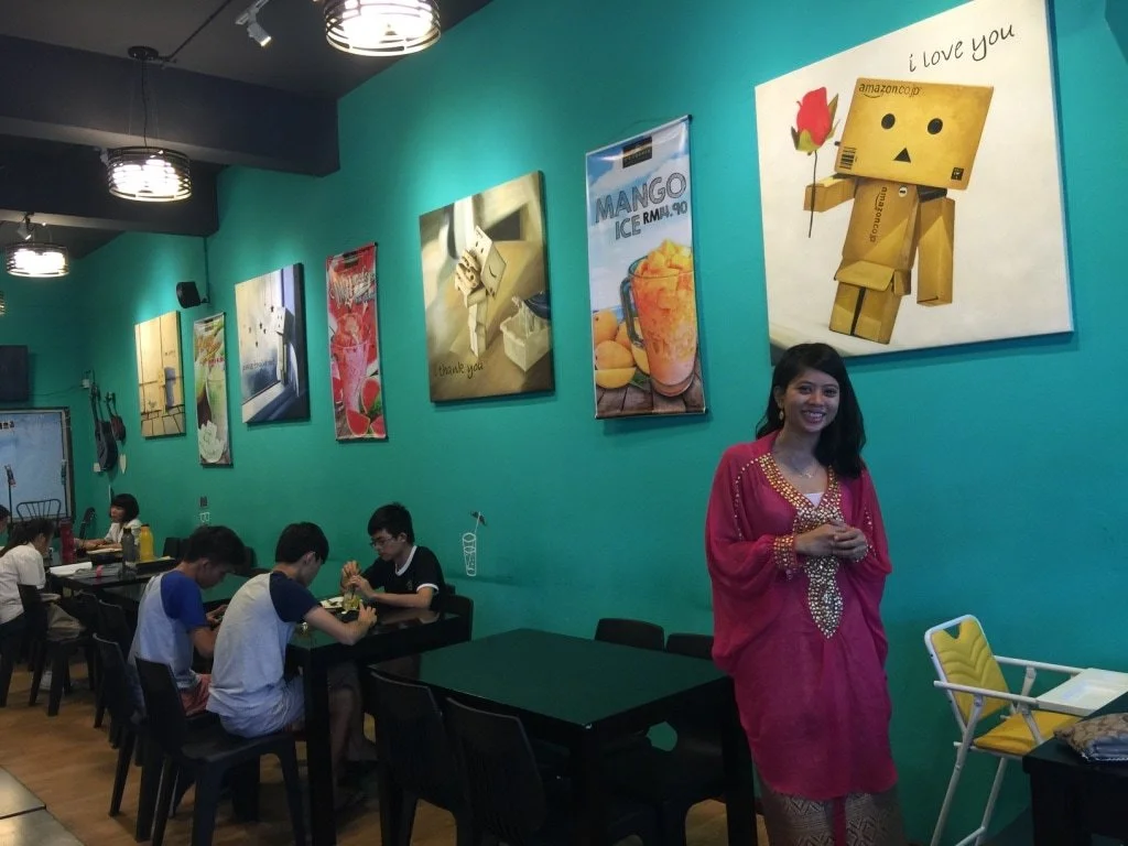 Young woman in pink traditional attire smiling inside a vibrant cafe with teal walls, artwork, and a group of young people dining at tables.