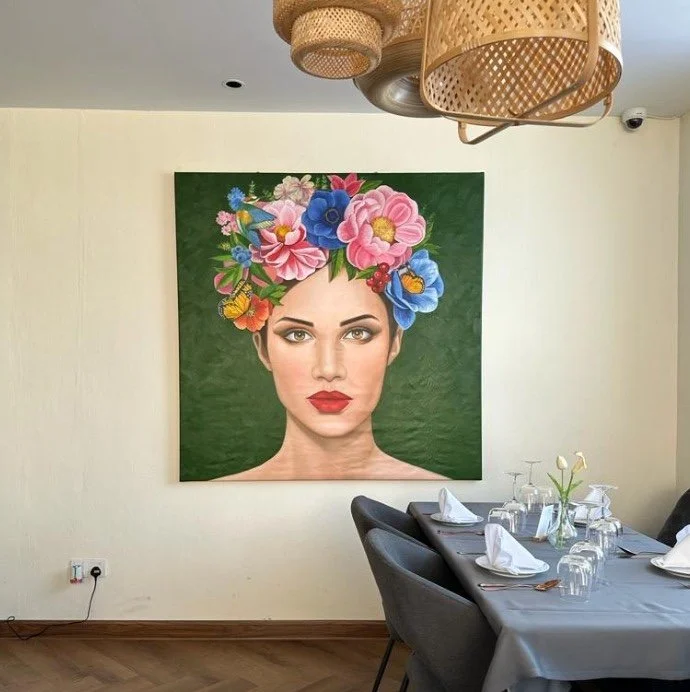 A large portrait of a woman with brown hair, red lipstick, and a floral crown of colorful flowers, hangs on a light-colored wall in a dining room with a table set with white napkins, glasses, and a small flower arrangement.
