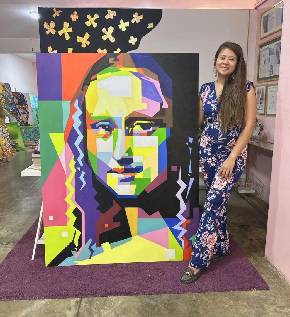 A woman in floral pajamas standing next to a colorful portrait of a woman, painted in a geometric, abstract style, displayed on an easel in an art gallery or store.