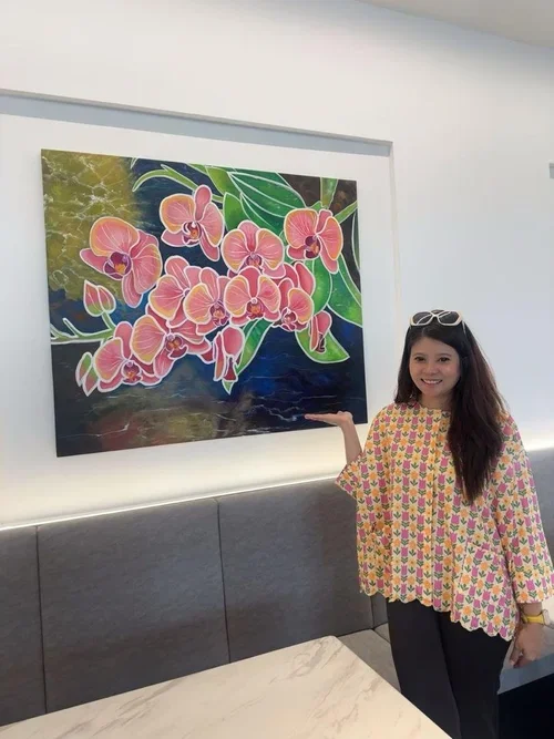A woman with long hair wearing glasses on her head, standing indoors, pointing at a colorful painting of pink and orange orchids on a dark background.