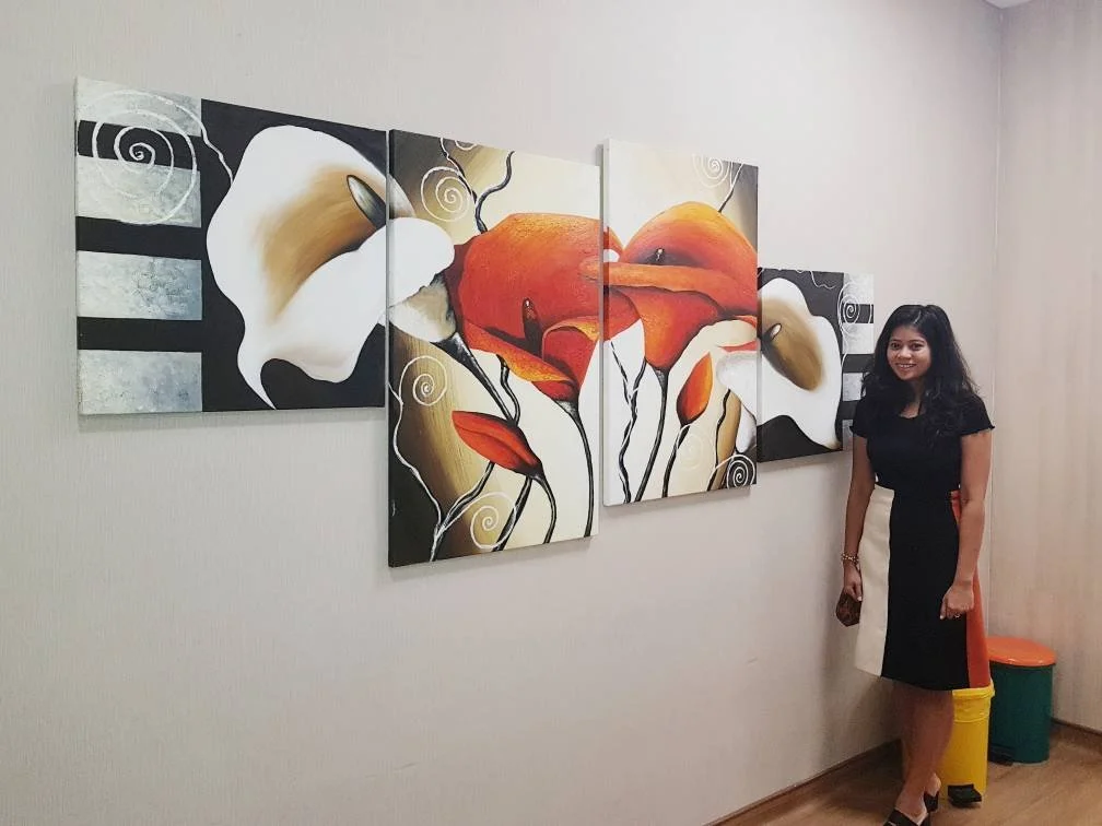 A woman standing next to a multi-panel floral painting of calla lilies and abstract flowers on a light-colored wall.