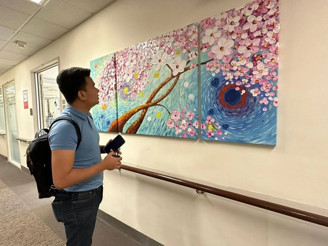 A teenage boy with a backpack looking at a multi-panel painting of a cherry blossom tree with swirling blue background and pink flowers.