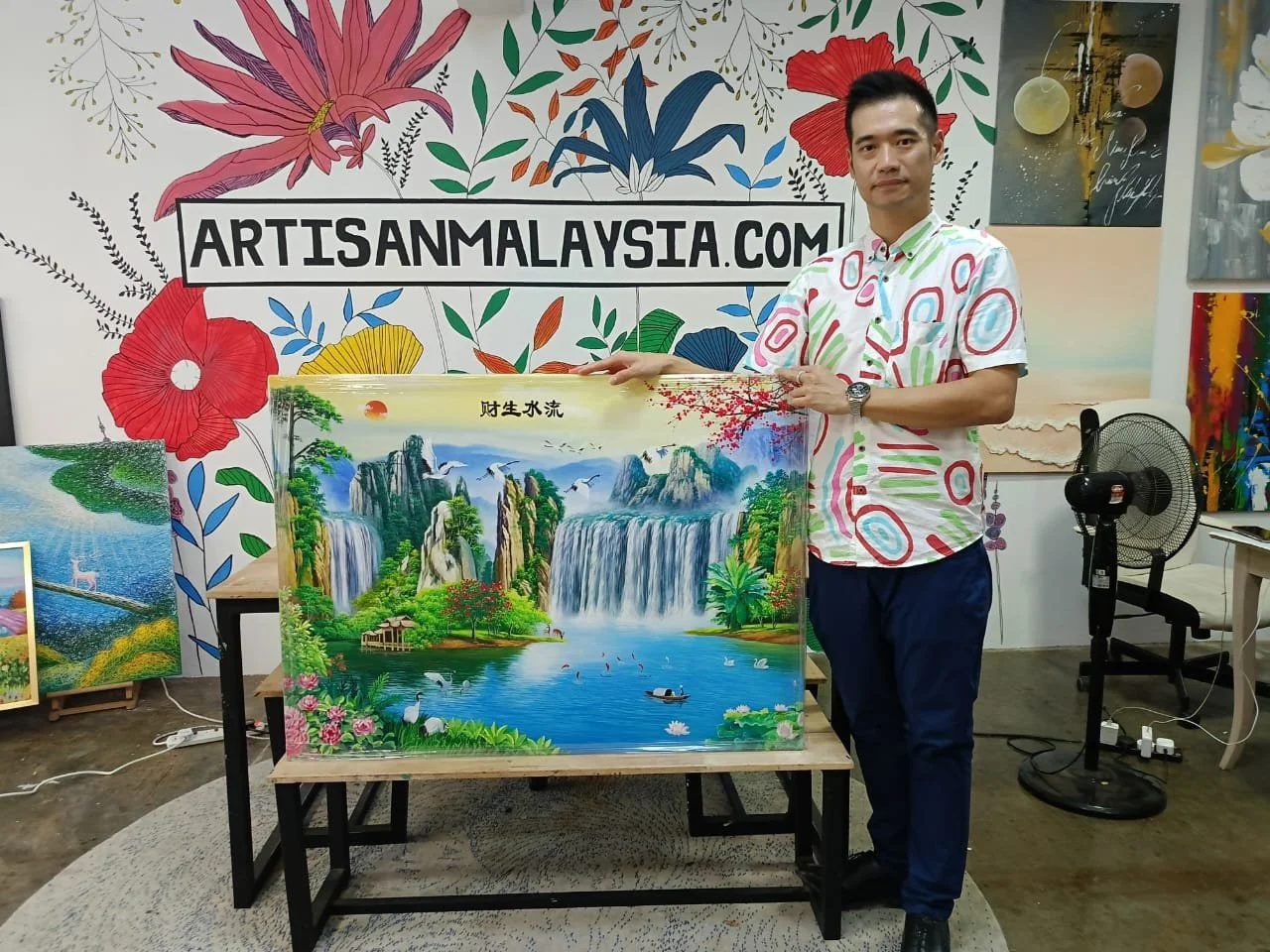 Man standing next to a colorful landscape painting featuring waterfalls, mountains, trees, swans, and a boat in a river setting.