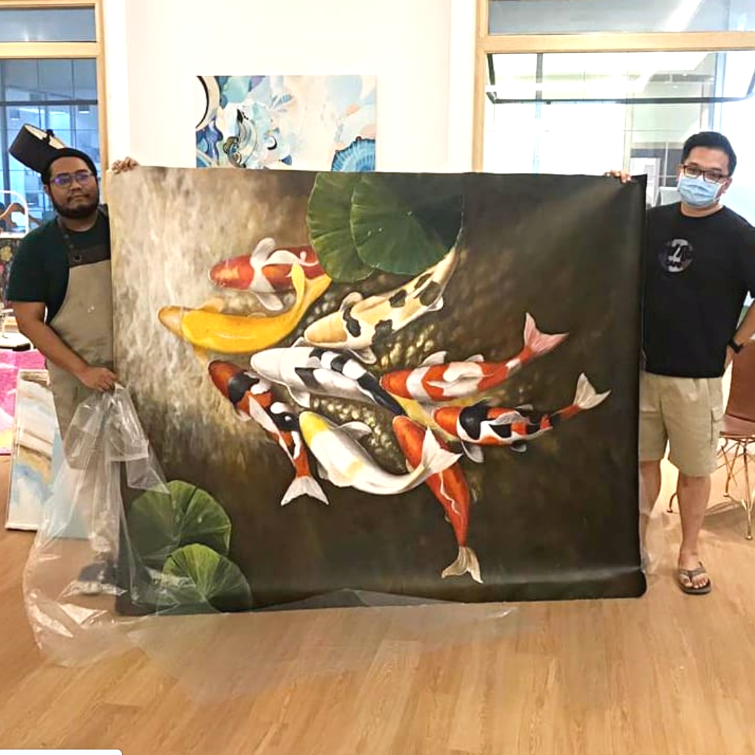 Two people, one wearing a face mask, hold a large colorful painting of fish and a large green leaf. The setting appears to be indoors, likely at an art exhibit or gallery.