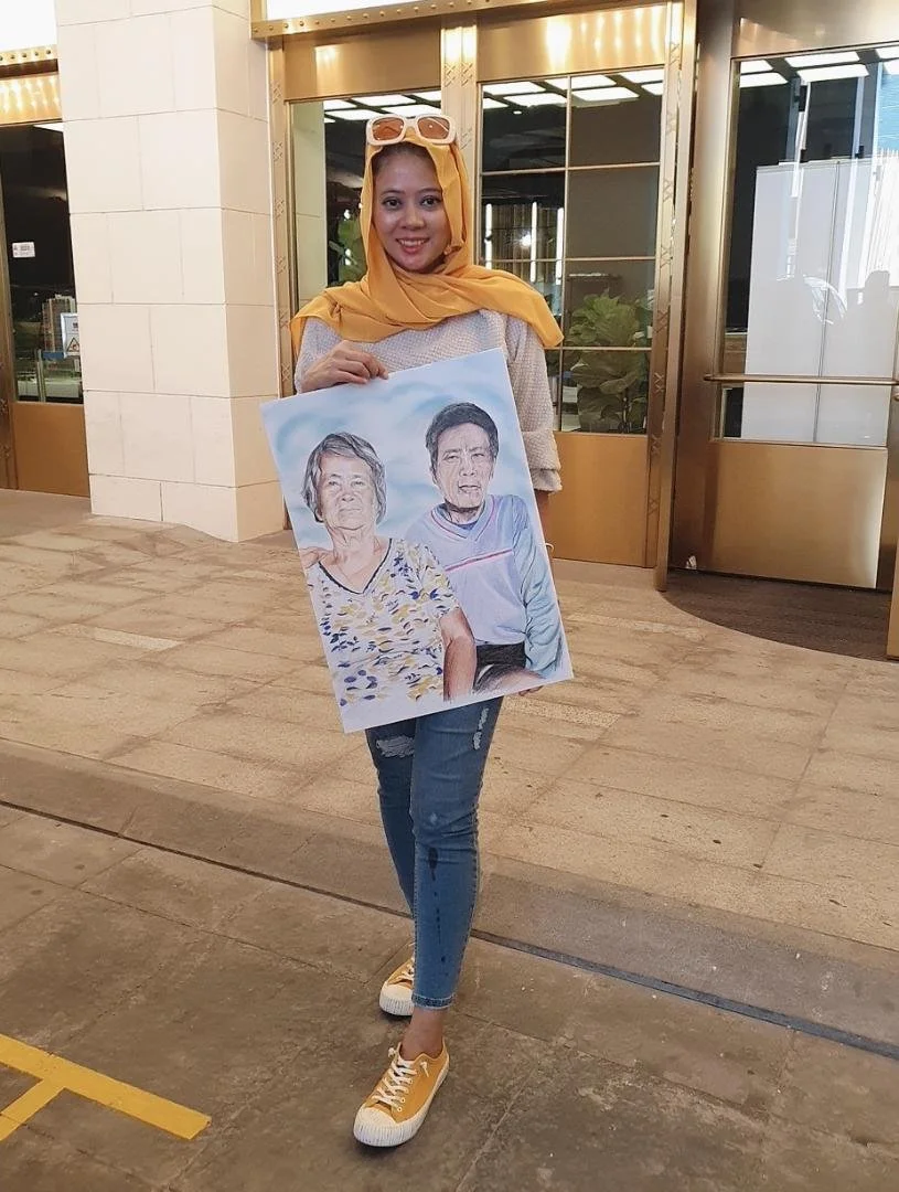 A woman in a yellow headscarf and sunglasses holding a watercolor portrait of an elderly couple. She is wearing ripped jeans, yellow sneakers, and is standing outside near a modern building with glass and gold accents.
