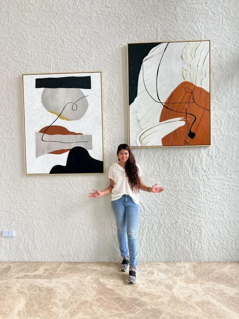 A young woman with long dark hair wearing a white shirt and blue jeans standing in front of a textured off-white wall with two large abstract art paintings. She is smiling and has her arms slightly extended.