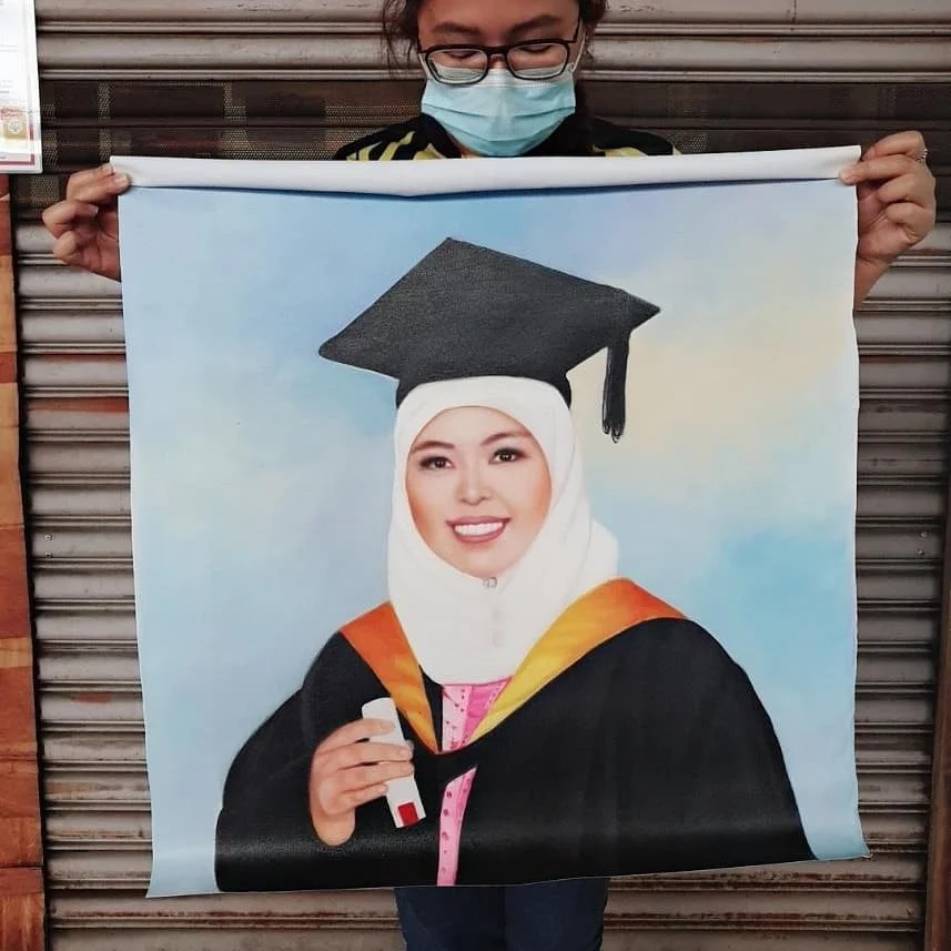 A person wearing glasses and a face mask holding a portrait of a woman in a graduation gown with a mortarboard, smiling and holding a diploma.
