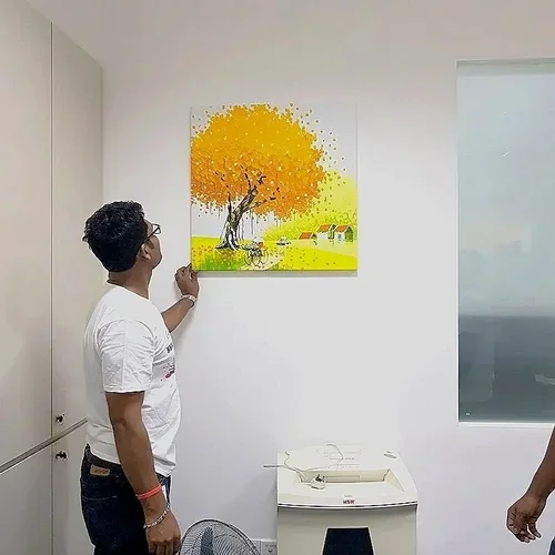 A man observing a painting of a vibrant orange and yellow tree with a small house and boat on a green field, hanging on a plain white wall.