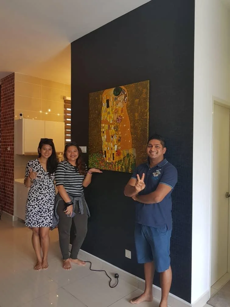 Three people standing indoors, smiling, posing next to a large framed artwork on a dark wall. The artwork features a colorful image of a couple embracing. The people are casually dressed, two women and one man, all barefoot.