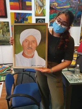 A woman wearing glasses and a face mask holding a framed portrait of an older man in traditional attire with a turban.