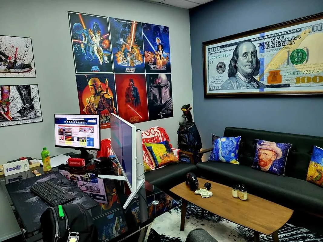 A room decorated with Star Wars posters, a large framed $100 bill artwork on the wall, a computer setup with dual monitors, a couch with colorful pillows, a coffee table with small bottles, and various electronic items around.