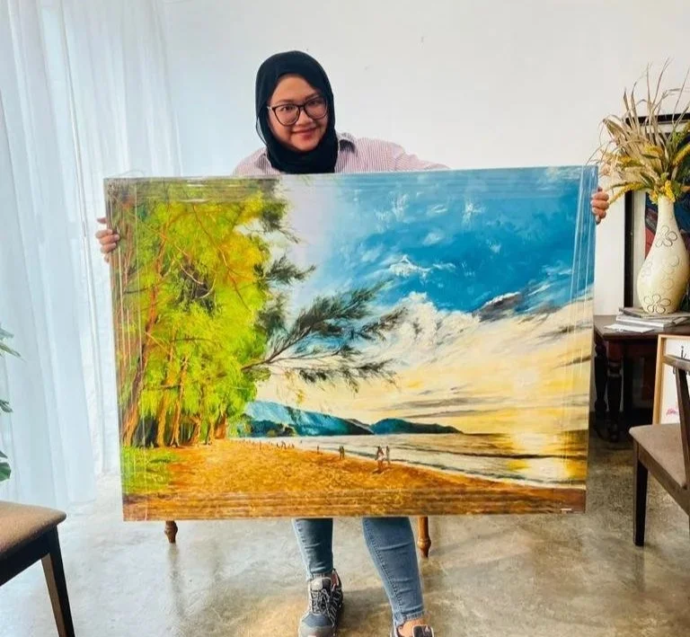 A woman wearing a black hijab and glasses is holding a large landscape painting of a beach with trees on the left and hills in the distance, under a partly cloudy sky.