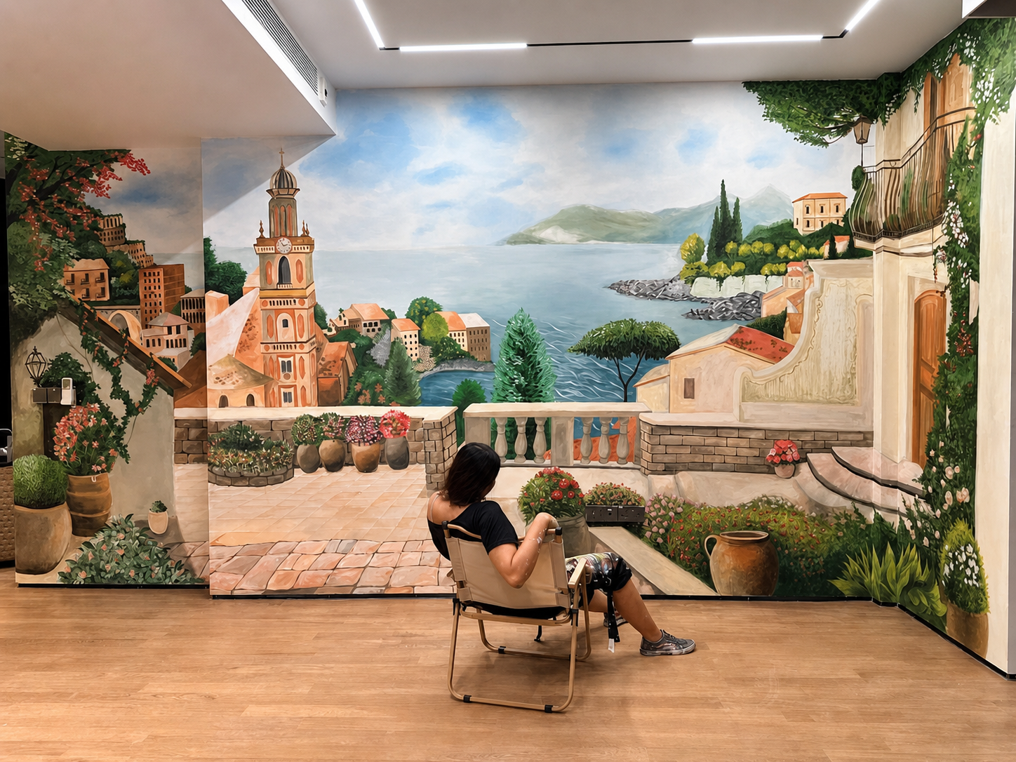 Person sitting in a chair in front of a mural depicting a scenic seaside village with a church tower, colorful buildings, trees, and mountains in the background.