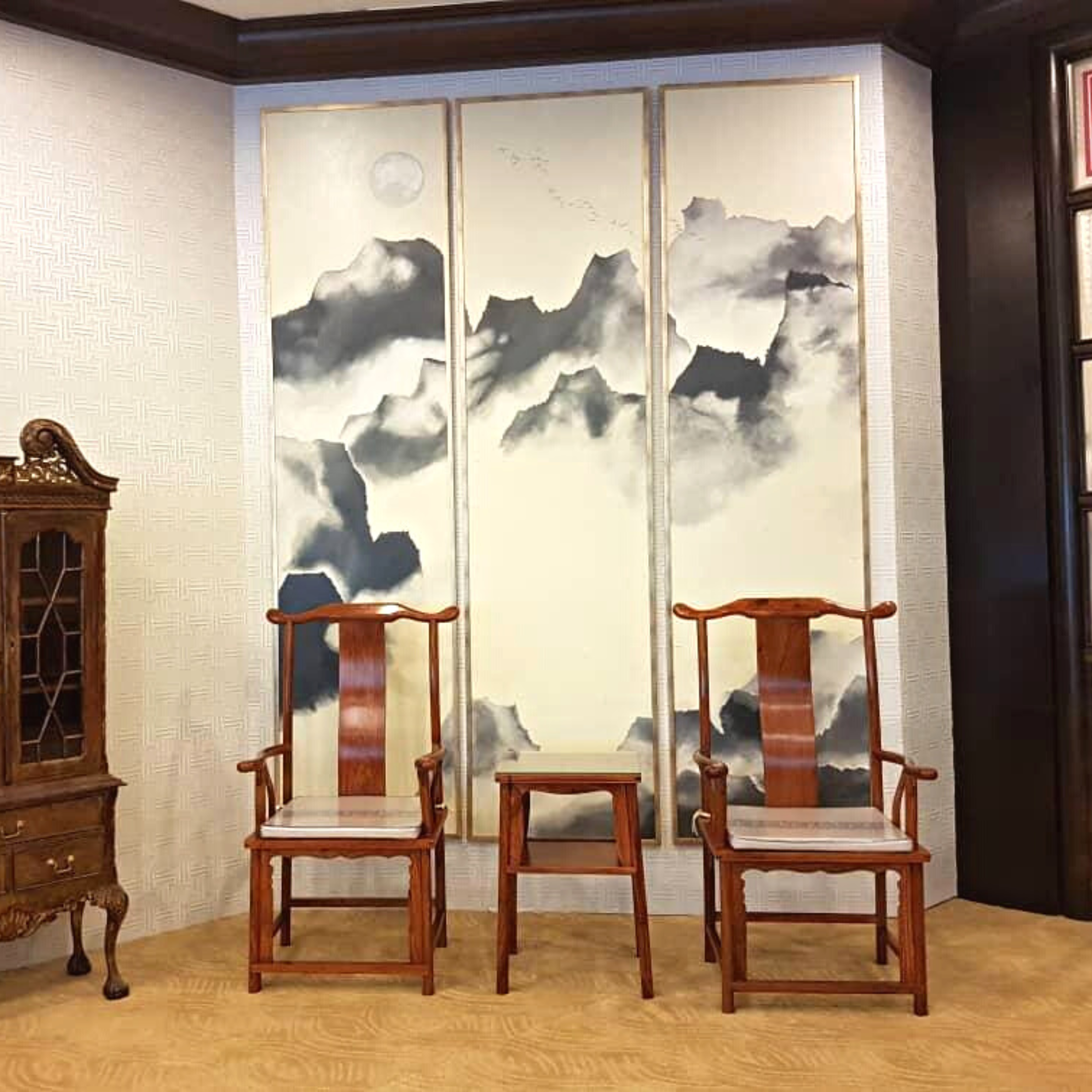 A large folding screen with a mountain landscape painting in gray tones, three panels connected with gold trim, set in a room with cream patterned wallpaper and wooden furniture.
