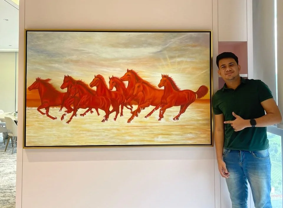 A man standing next to a large painting of several running orange horses on a beach at sunset.