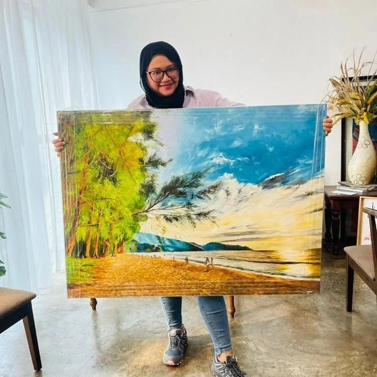 A woman wearing glasses, a black hijab, a pink striped shirt, and jeans is holding a large landscape painting of a beach scene with trees, water, and a cloudy sky.