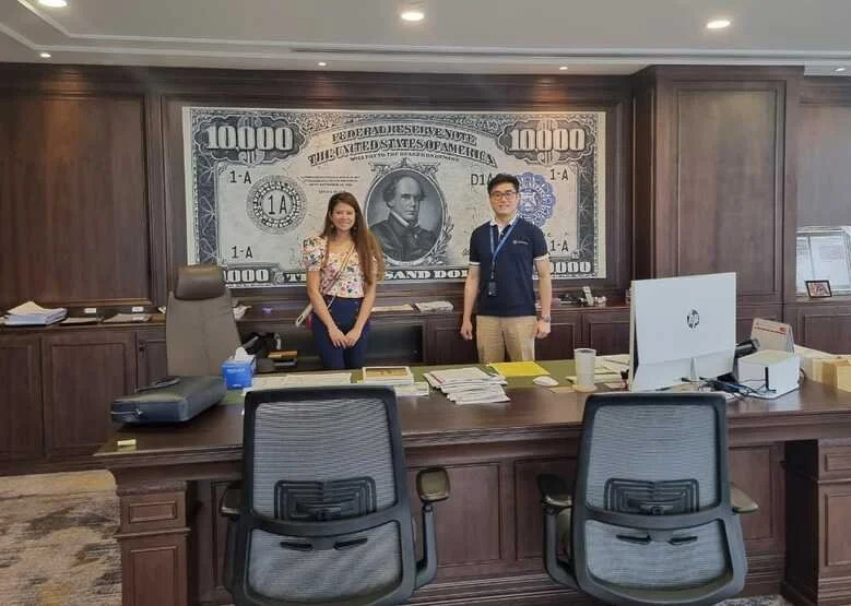 Two people standing in front of a large mural of a $10,000 bill with Benjamin Franklin's portrait in an office setting.