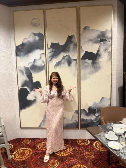 A woman in a pink lace dress standing in front of a large three-panel Asian-inspired ink wash mountain landscape painting with a moon and birds, inside a room with patterned carpet and a table set with dishes.