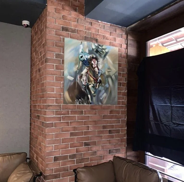 Painting of a cowboy riding a bucking horse on a brick wall in a room with leather seating and a window.