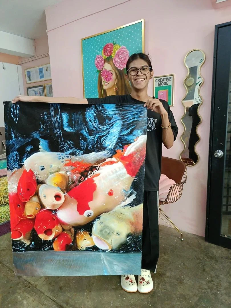 Young woman holding a vibrant painting of colorful koi fish swimming in water, smiling in a decorated room with pink walls, a mirror, and framed artwork.