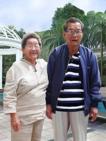 An elderly woman and man standing outdoors near a pool with lush green trees in the background. They are smiling, dressed casually, and appear to be enjoying a sunny day.