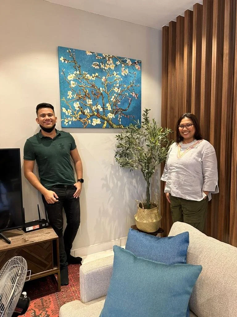 Two people, a man and a woman, standing in a living room. The man is on the left, wearing a dark green polo shirt and dark pants. The woman is on the right, wearing a white top and green pants. There is a painting of a blossoming tree on the wall beh