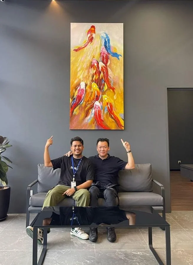 Two men sitting on a gray couch in a modern lobby, smiling and pointing upward with their index fingers. Behind them hangs a large colorful abstract painting of koi fish on a dark gray wall. A glass coffee table is in front of them, and a potted plant is on the left.