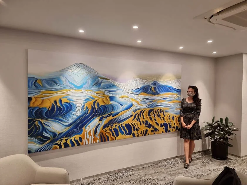 A woman wearing a face mask and a black dress standing next to a colorful abstract landscape painting of mountains and hills in shades of blue, yellow, and white, mounted on a white gallery wall in an indoor setting.