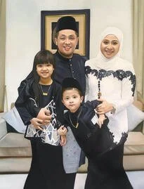 A family of four posing together indoors. The father is wearing a traditional black hat and dark clothing, the mother is dressed in a white headscarf and white top, and two children, a girl and a boy, are standing in front. The girl has a dark jacket