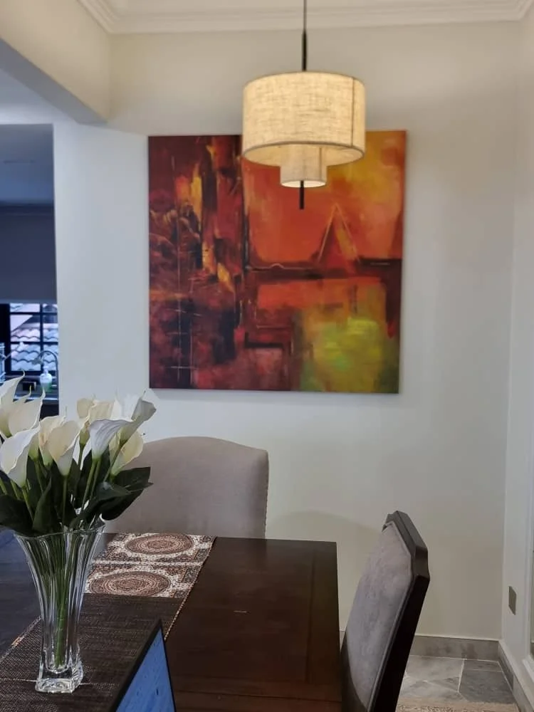A dining room with a dark wood table, a beige upholstered chair, and a glass vase with white calla lilies. An abstract painting with warm hues of orange, red, and yellow hangs on the wall, illuminated by a hanging beige lampshade.