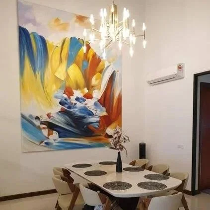 A dining room with a round table, six chairs with beige cushions, and a black vase with dried flowers on the table. An abstract painting with vibrant colors hangs on the wall, and a modern chandelier with multiple lights hangs above the table. An air conditioner is mounted on the wall.