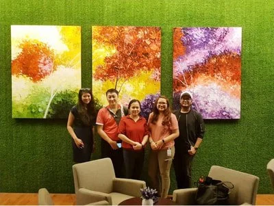 Five people standing in front of three colorful, abstract tree paintings on a green wall in an indoor setting.