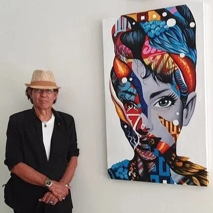 A woman wearing a beige hat and black jacket stands next to a colorful abstract portrait of a woman on a white wall.