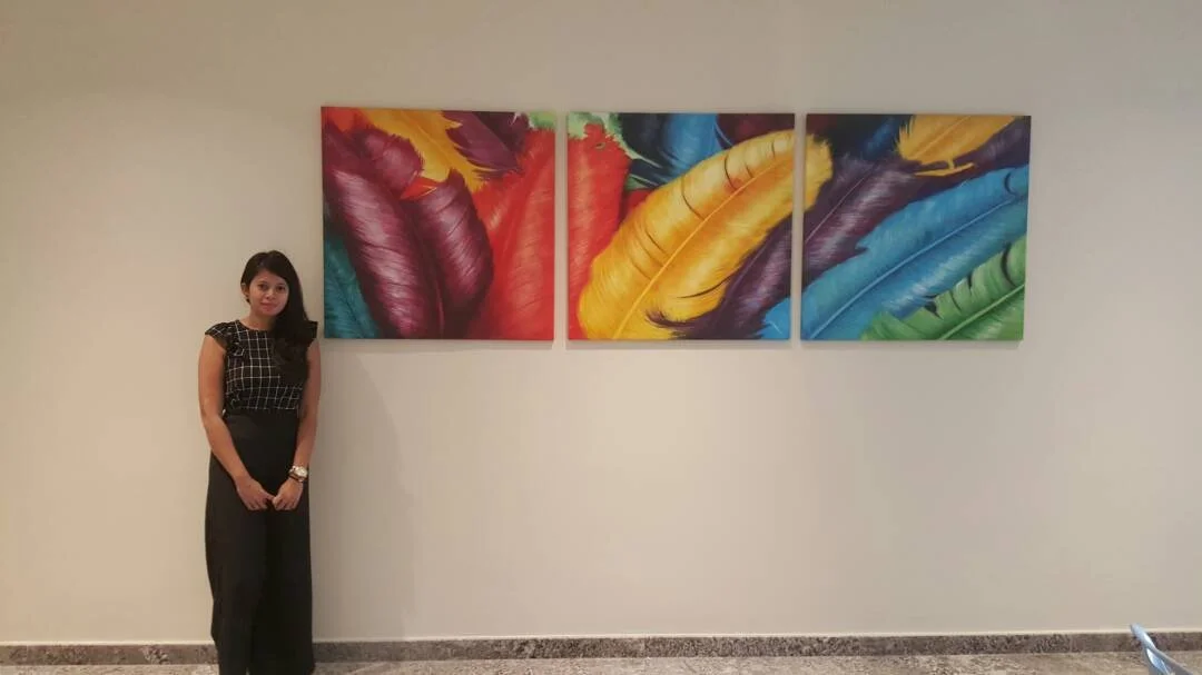 A woman standing next to a triptych of colorful feather paintings on a plain wall.