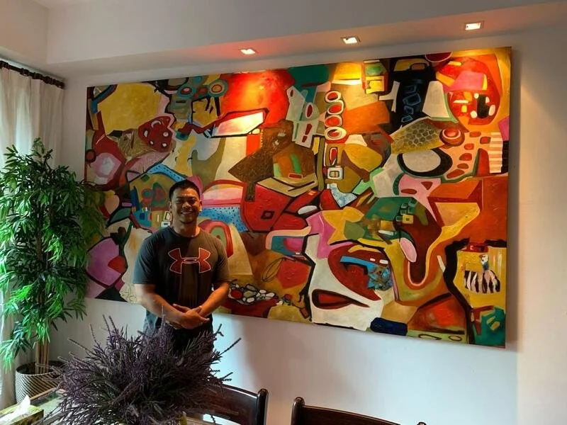 A man standing in front of an abstract colorful painting in a room with a potted plant on the left and a window with white curtains.