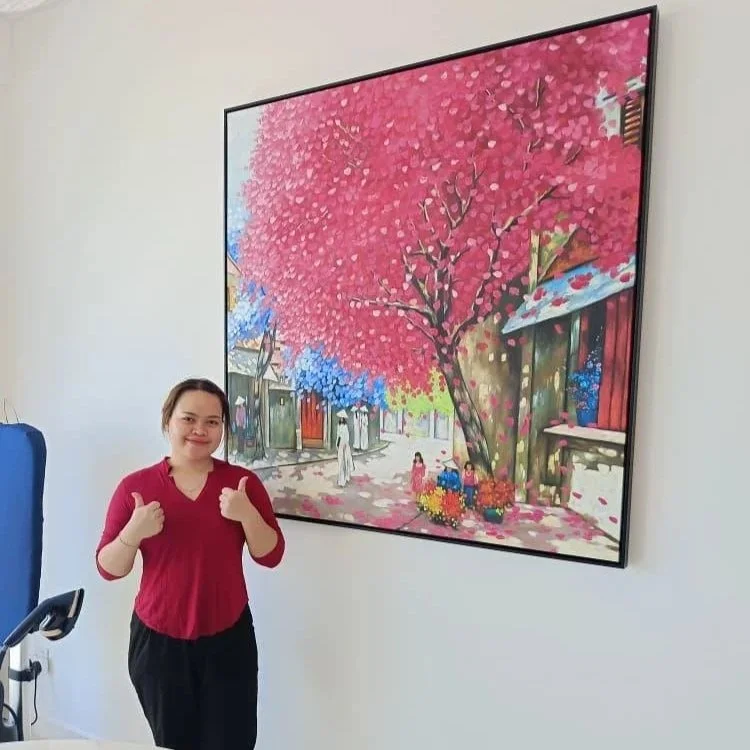 A woman in a red top giving a thumbs up next to a large colorful painting of a street scene with pink and blue trees and houses.