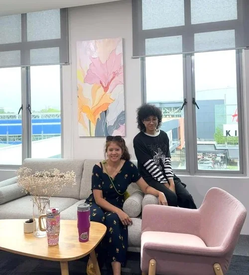 Two young women sitting in a modern lounge area with large windows. One is seated on a sofa, smiling, while the other is standing behind her. There is a wooden coffee table with drinks and a bouquet of flowers, and a pink armchair is in the foregroun