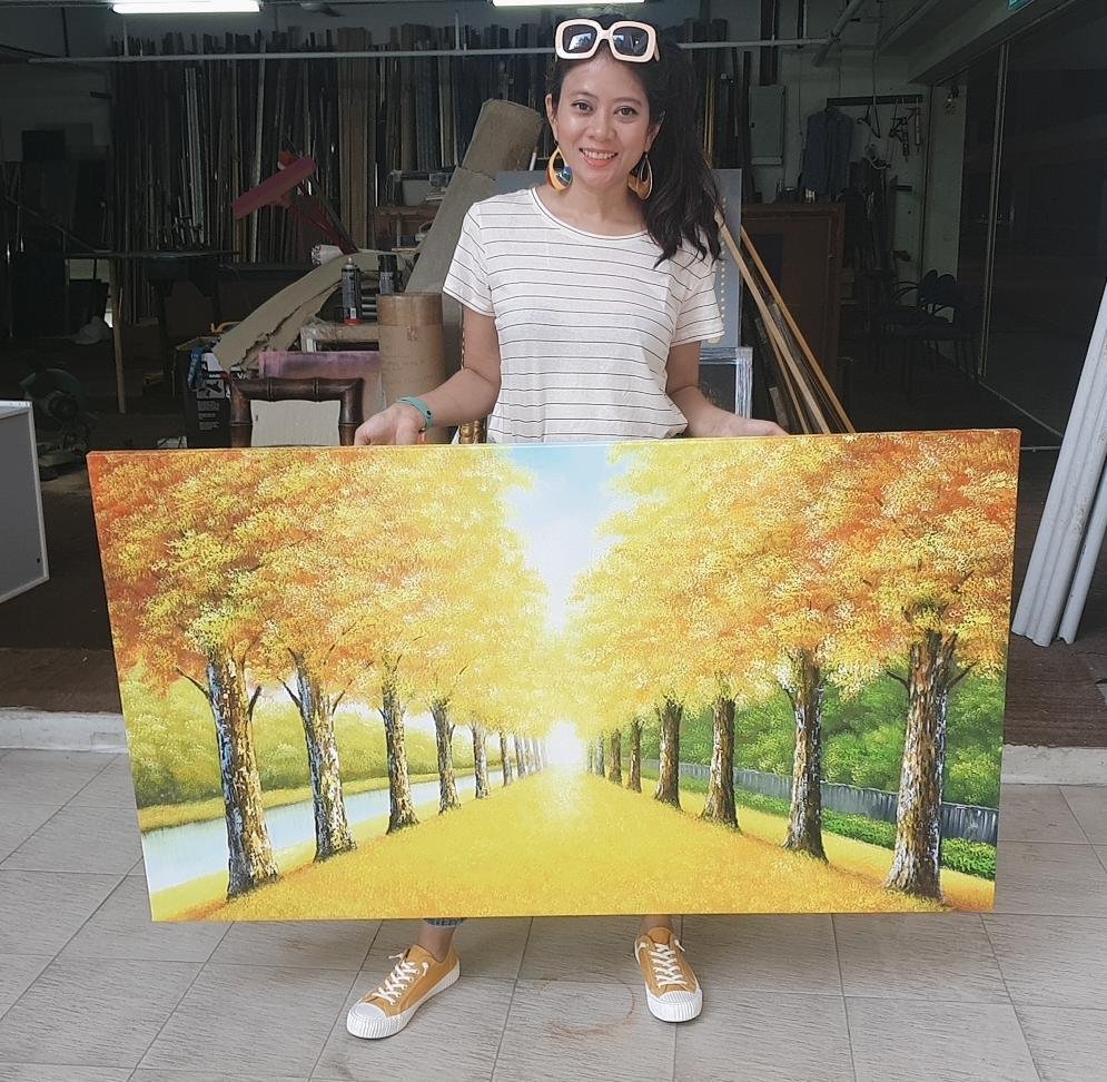 Woman holding a large painting of a tree-lined pathway with fall foliage.