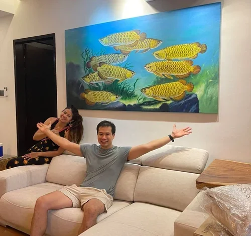 Two people sitting on a beige sofa in a living room, smiling, with a large colorful painting of fish swimming underwater hanging on the wall behind them.