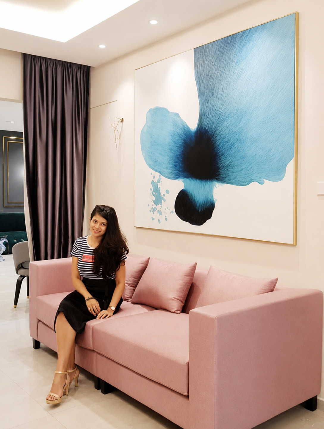 A woman sitting on a pink sofa in a modern hotel lobby, smiling at the camera, with a large abstract blue flower painting on the wall behind her.